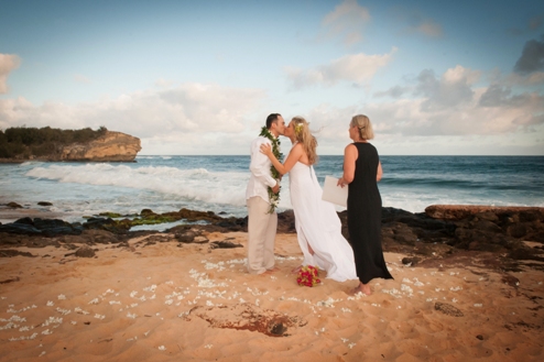 A bride and groom are kissing on a sandy beach while an officiant in a black dress looks on, with the ocean and cliffs in the background.