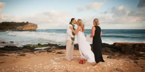 A bride and groom are kissing on a sandy beach while an officiant in a black dress looks on, with the ocean and cliffs in the background.