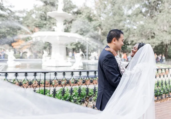 A bride and groom smiling at each other in a park setting with a white fountain in the background.