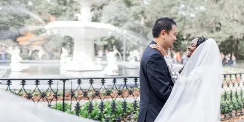 A bride and groom smiling at each other in a park setting with a white fountain in the background.