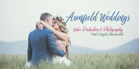 A bride and groom embracing in a grassy field with the Aronfield Weddings logo and text overlay.