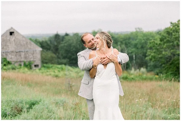 A bride and groom embracing in a grassy field with a rustic wooden barn in the background.