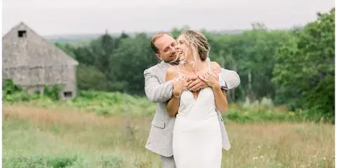A bride and groom embracing in a grassy field with a rustic wooden barn in the background.