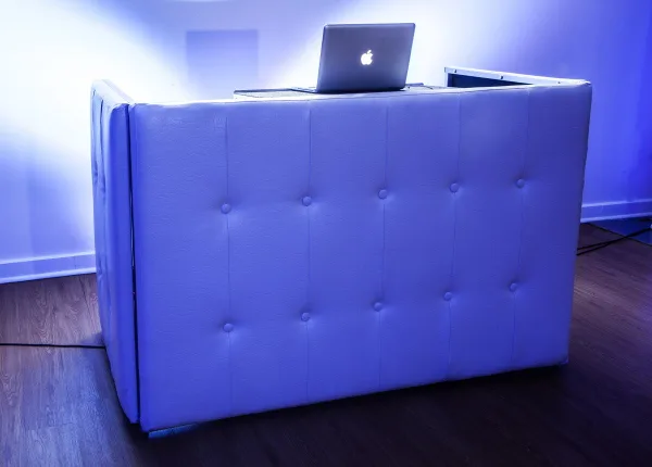 A white tufted DJ booth with a laptop on top, illuminated by blue and purple lighting.