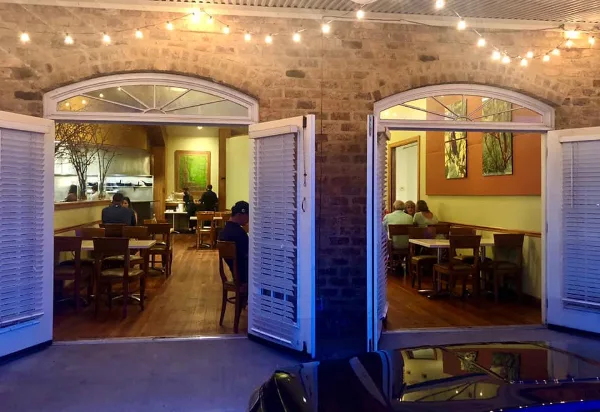 Interior view of a restaurant dining area with wooden tables, chairs, and an open kitchen visible through arched doorways.