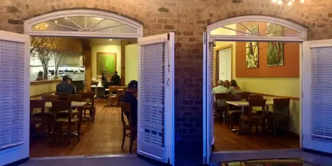 Interior view of a restaurant dining area with wooden tables, chairs, and an open kitchen visible through arched doorways.