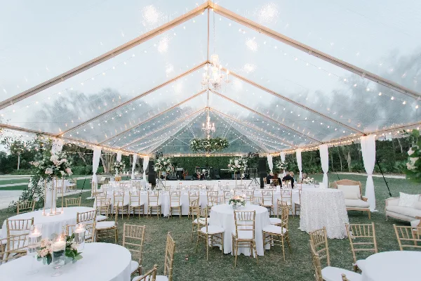 An outdoor wedding reception setup inside a clear tent with white table linens, gold chairs, and string lighting.
