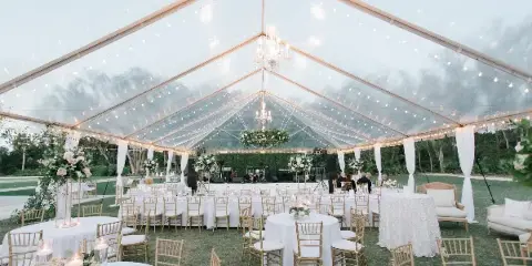 An outdoor wedding reception setup inside a clear tent with white table linens, gold chairs, and string lighting.
