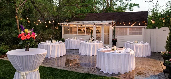 An outdoor event setup featuring multiple round tables with white linens and a mobile bar under string lights.