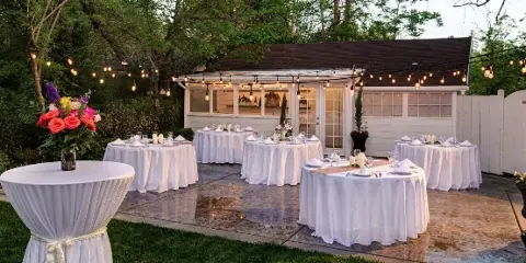 An outdoor event setup featuring multiple round tables with white linens and a mobile bar under string lights.