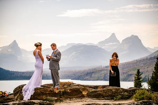 A bride and groom stand on a rocky overlook with a female officiant holding a folder during a wedding ceremony, with mountains and a lake in the background.