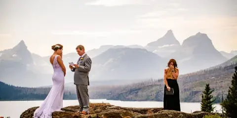 A bride and groom stand on a rocky overlook with a female officiant holding a folder during a wedding ceremony, with mountains and a lake in the background.