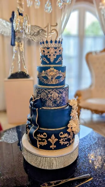 A tall, multi-tiered navy blue cake decorated with ornate gold patterns, a crown topper, and floral accents, displayed on a glittering stand.