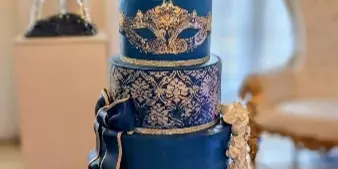 A tall, multi-tiered navy blue cake decorated with ornate gold patterns, a crown topper, and floral accents, displayed on a glittering stand.
