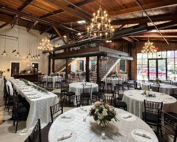 Interior of a wedding venue with round tables, white tablecloths, floral centerpieces, and hanging chandeliers.