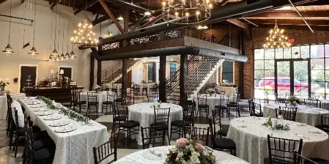 Interior of a wedding venue with round tables, white tablecloths, floral centerpieces, and hanging chandeliers.