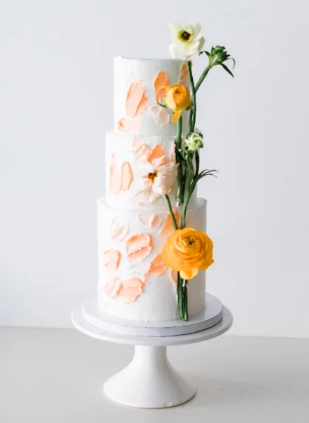 A three-tier white wedding cake decorated with peach-colored brushstrokes and fresh yellow and white flowers on a white cake stand.