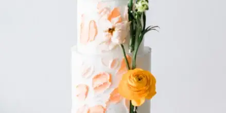 A three-tier white wedding cake decorated with peach-colored brushstrokes and fresh yellow and white flowers on a white cake stand.