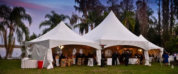 Large white event tent set up outdoors with tables, chairs, and guests at an evening reception.