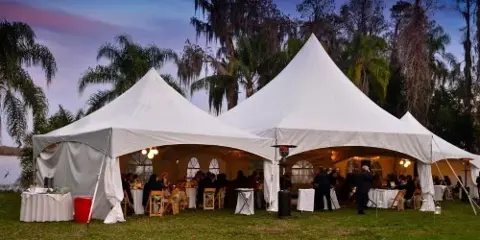 Large white event tent set up outdoors with tables, chairs, and guests at an evening reception.