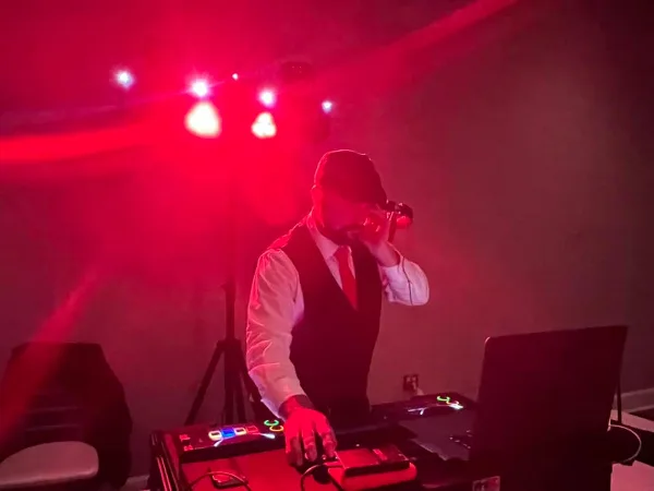 A professional DJ in a vest and tie performing at an event under red stage lighting.