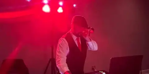 A professional DJ in a vest and tie performing at an event under red stage lighting.