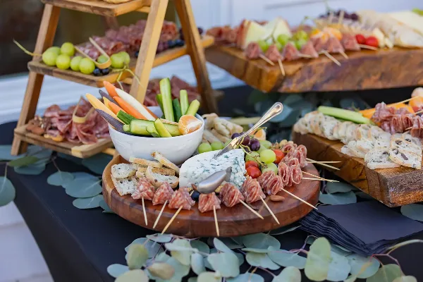 A catering table setup featuring a tiered wooden stand with charcuterie, fresh vegetables, and breads.
