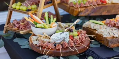 A catering table setup featuring a tiered wooden stand with charcuterie, fresh vegetables, and breads.