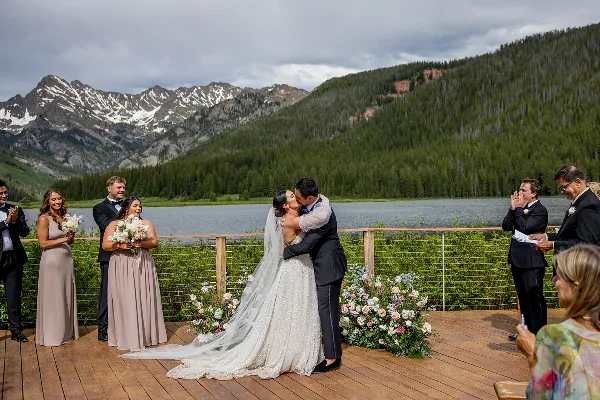 A bride and groom sharing a kiss during a wedding ceremony on a wooden deck overlooking a mountain lake.