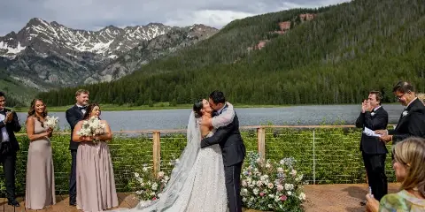 A bride and groom sharing a kiss during a wedding ceremony on a wooden deck overlooking a mountain lake.