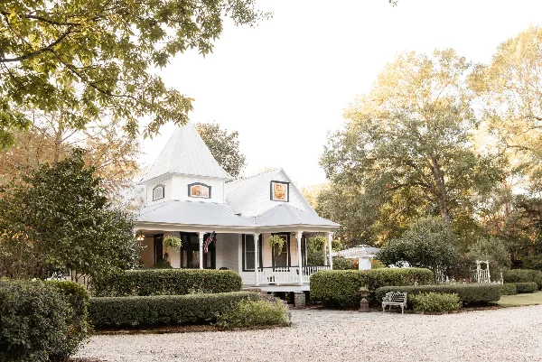 Exterior view of a white Victorian-style house with a conical roof, surrounded by manicured hedges, trees, and a gravel courtyard.