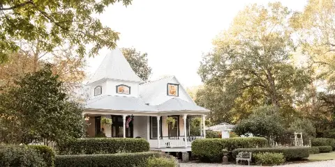 Exterior view of a white Victorian-style house with a conical roof, surrounded by manicured hedges, trees, and a gravel courtyard.