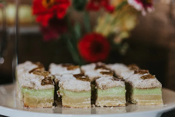 A plate of layered, square-cut pastries with green and yellow layers, topped with powdered sugar and nuts, set against a blurred background of red flowers.