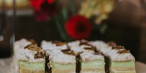 A plate of layered, square-cut pastries with green and yellow layers, topped with powdered sugar and nuts, set against a blurred background of red flowers.