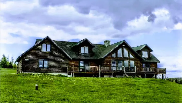 Exterior view of a large rustic log lodge with a green roof and wooden deck situated on a grassy hillside.