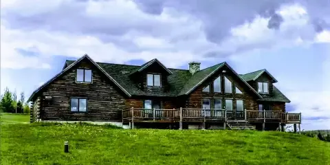 Exterior view of a large rustic log lodge with a green roof and wooden deck situated on a grassy hillside.