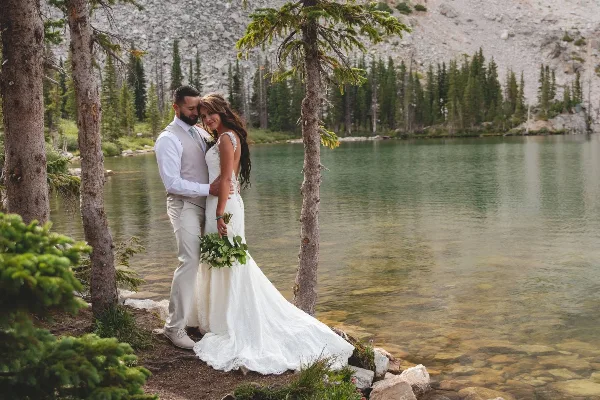 A bride and groom standing together by a mountain lake with evergreen trees in the background.