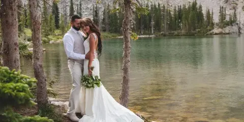 A bride and groom standing together by a mountain lake with evergreen trees in the background.