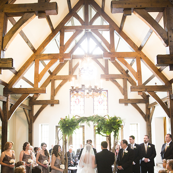 Interior view of a historic castle hall with exposed wooden beams and a stained-glass window during a wedding ceremony.