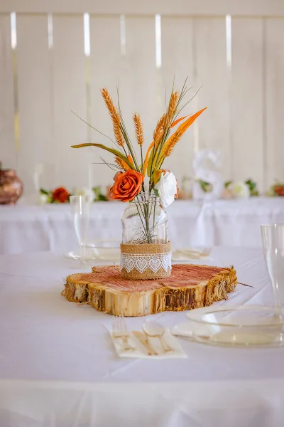 A rustic wedding table setting featuring a mason jar centerpiece with wheat and roses on a wood slice, set against a white barn interior.