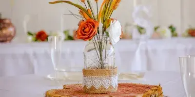 A rustic wedding table setting featuring a mason jar centerpiece with wheat and roses on a wood slice, set against a white barn interior.