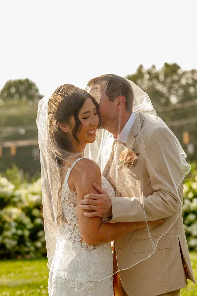 A bride and groom embracing and kissing outdoors, with the bride wearing a lace wedding dress and veil.