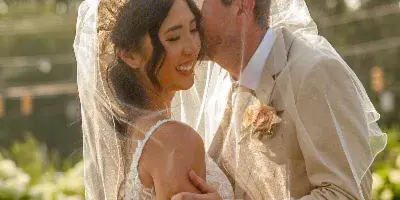 A bride and groom embracing and kissing outdoors, with the bride wearing a lace wedding dress and veil.