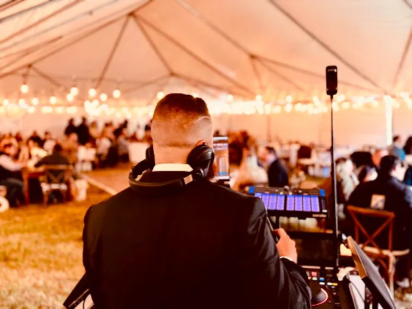 A DJ in a suit with headphones on, operating a Pioneer DJ controller at a wedding reception under a tent with string lights and guests in the background.