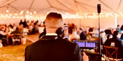 A DJ in a suit with headphones on, operating a Pioneer DJ controller at a wedding reception under a tent with string lights and guests in the background.