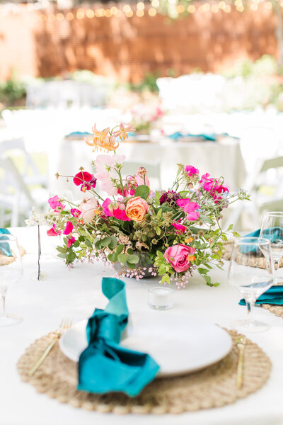 A close-up of a wedding table setting featuring a vibrant floral arrangement with pink and orange roses, white plates, and teal napkins on a white tablecloth.