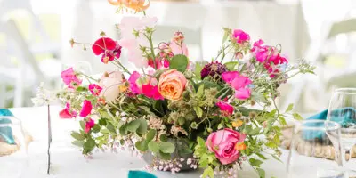 A close-up of a wedding table setting featuring a vibrant floral arrangement with pink and orange roses, white plates, and teal napkins on a white tablecloth.