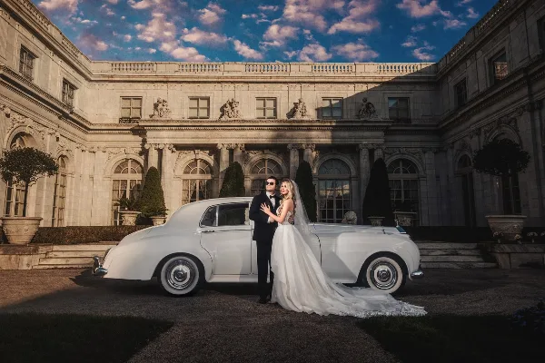 A bride and groom posing in front of a white vintage car at a grand stone estate.