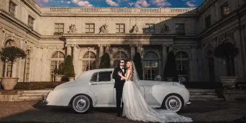 A bride and groom posing in front of a white vintage car at a grand stone estate.