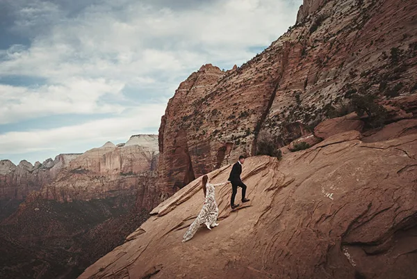 A bride and groom walking hand-in-hand on a rocky mountain slope with a vast canyon landscape in the background.